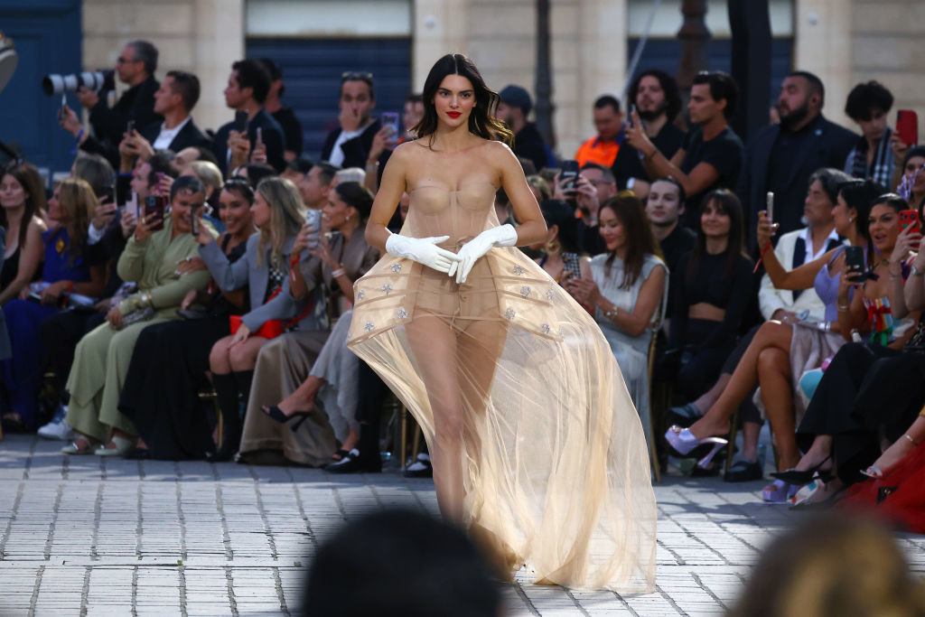 Kendall Jenner walking the runway in a sheer, strapless gown with gloves at a fashion show, surrounded by a seated audience taking photos and videos