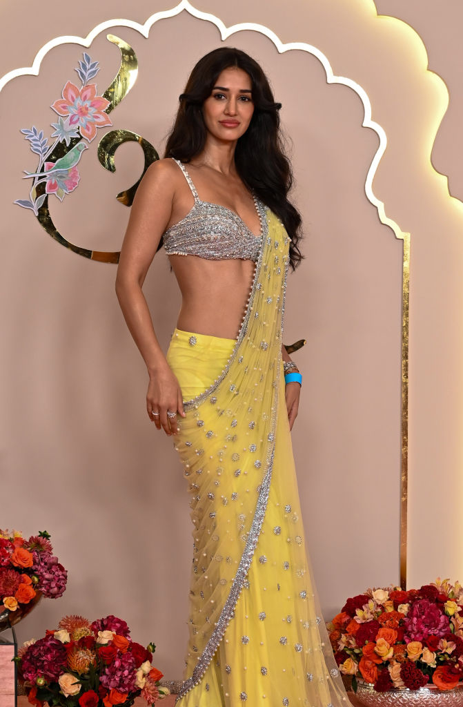 Disha wearing a sparkling bralette and a sari adorned with floral designs, surrounded by vases of colorful flowers
