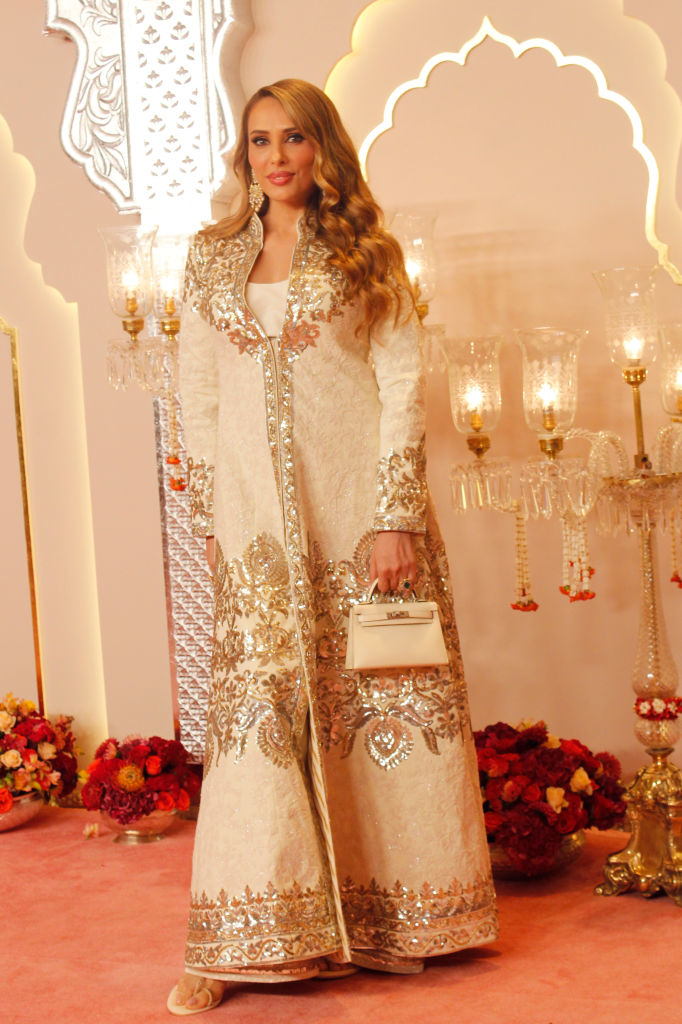 Iulia VĂąntur wearing an ornate, long coat dress holds a small handbag. The background features decorative lighting and flowers