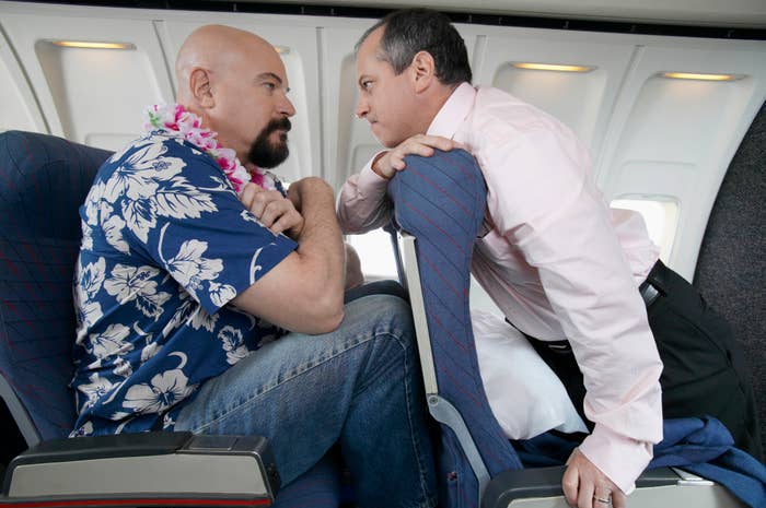 Two men, one in a floral shirt and lei, the other in a suit, sit in airplane seats facing each other intensely