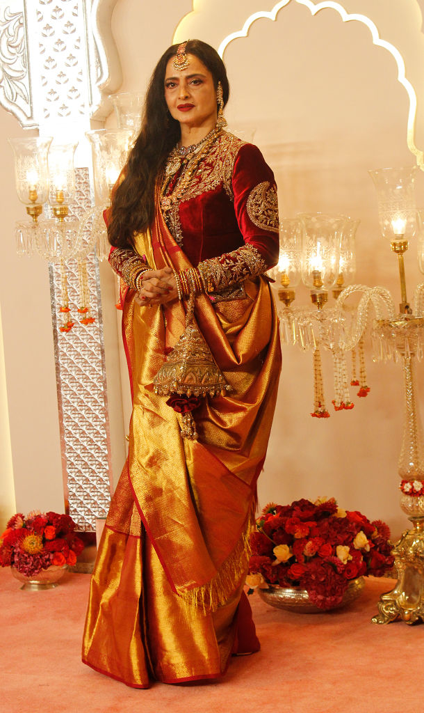 Rekha stands on a red carpet in an opulent sari with intricate embroidery, accessorized with traditional jewelry