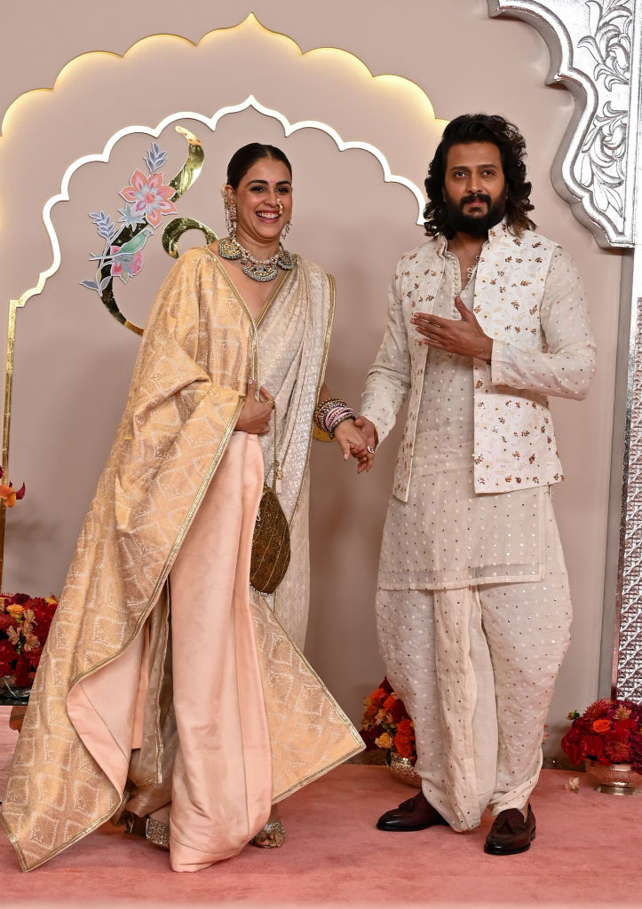 Riteish Deshmukh and Genelia D'Souza pose together in traditional attire on a decorated background. Riteish wears an embroidered kurta set, and Genelia wears a saree with jewelry