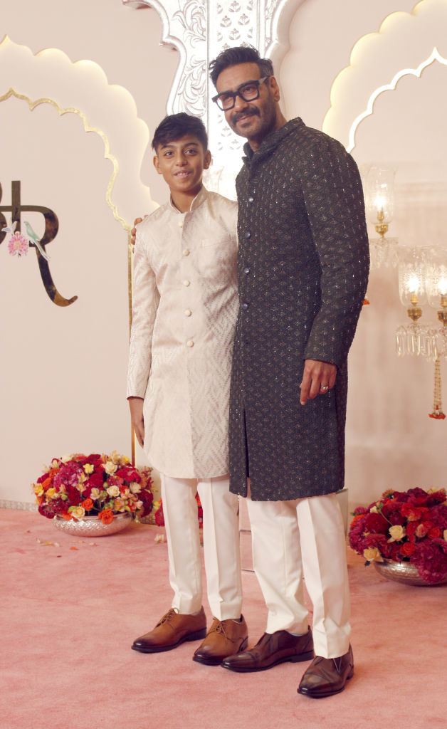 Ajay Devgn and his son, Yug Devgn, stand on a red carpet, both dressed in traditional Indian outfits. Ajay is in a dark sherwani, and Yug is in a light-colored one