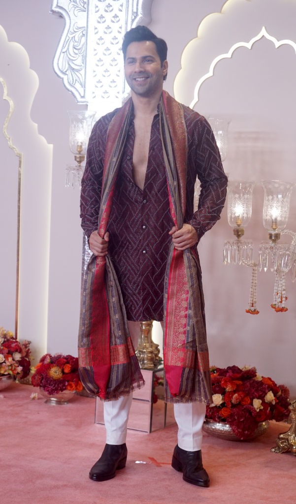 Varun Dhawan poses on the red carpet in traditional attire with a richly patterned shawl and neutral pants