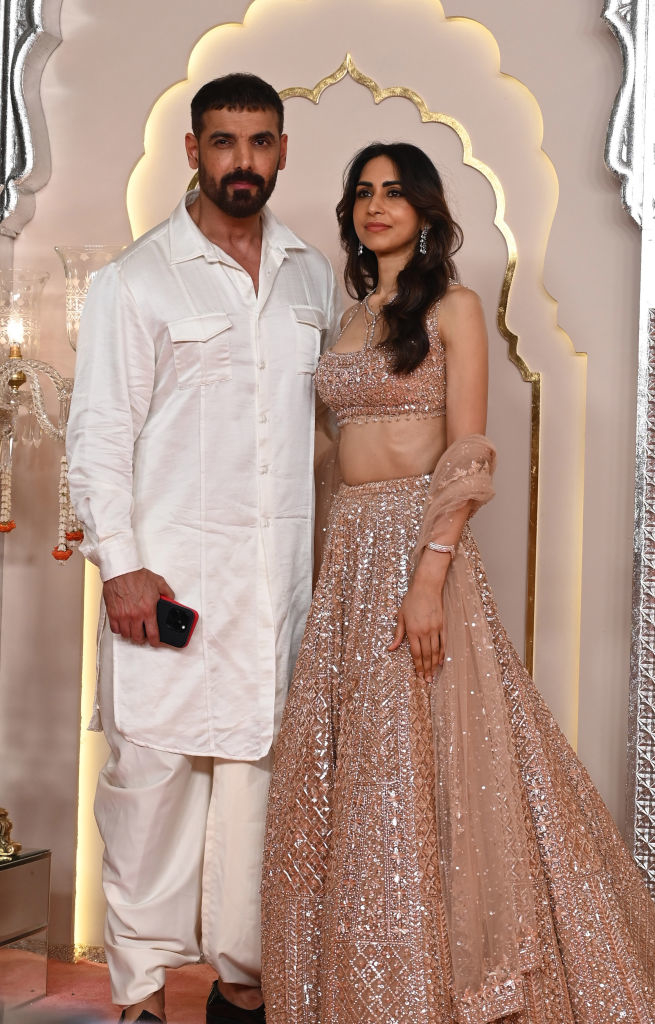 John Abraham and Priya Runchal pose on a red carpet, with John in a kurta and trousers and Priya in a sparkling traditional lehenga