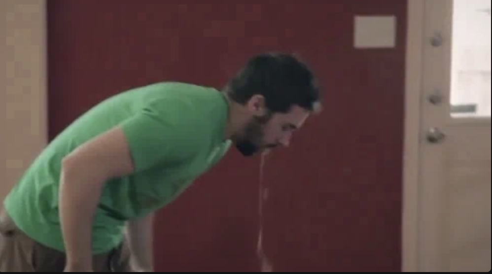 A man in a green shirt is bent over, vomiting. The background is indoors with visible portions of a red wall and a white door