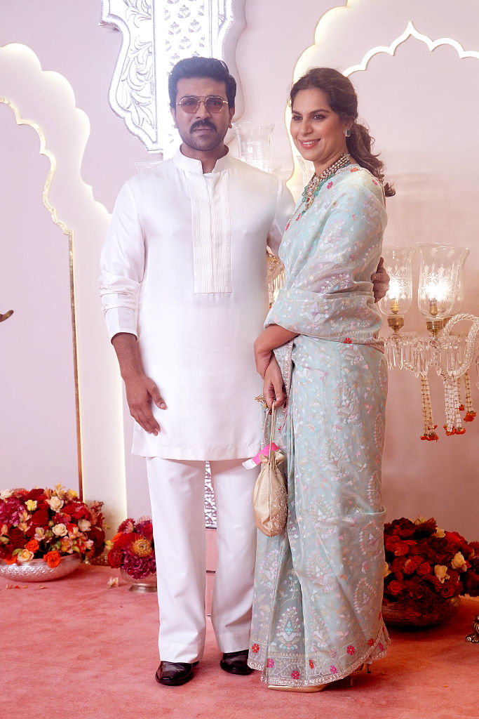 Ram Charan and Upasana Kamineni dressed in traditional attire at an event; Ram in a light kurta and Upasana in a light saree holding a small bag