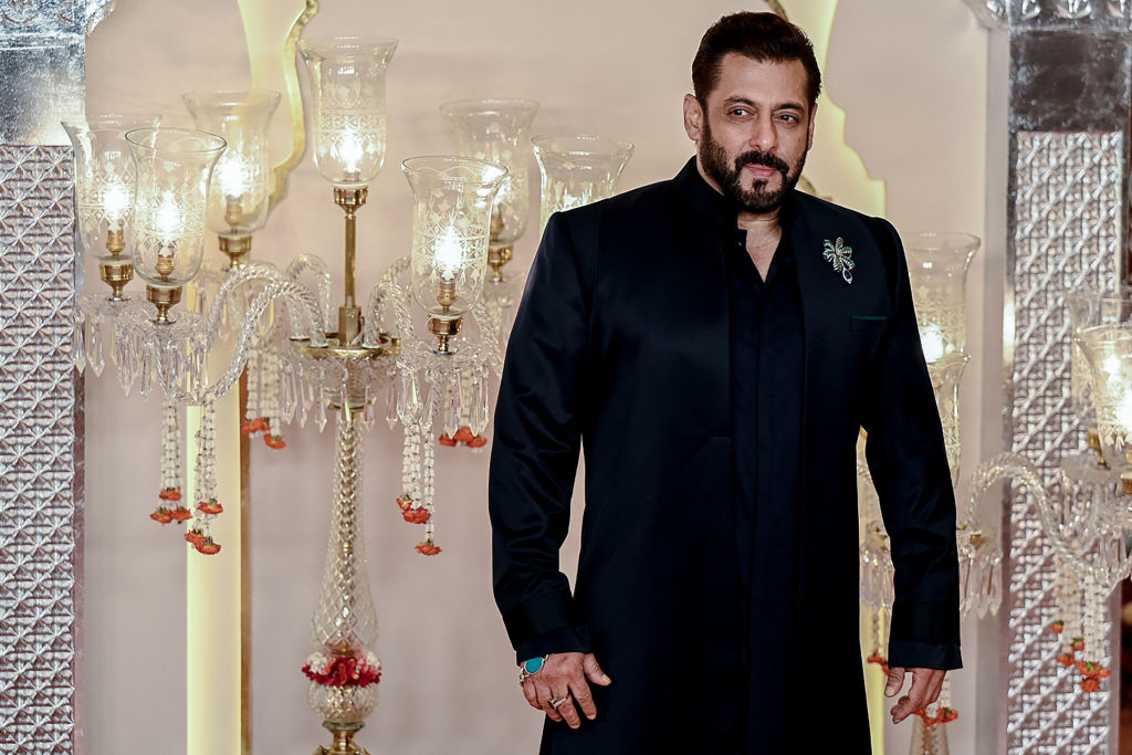 Salman Khan poses on a red carpet, wearing a stylish traditional outfit adorned with a brooch. Elegant candlestick lamps are in the background