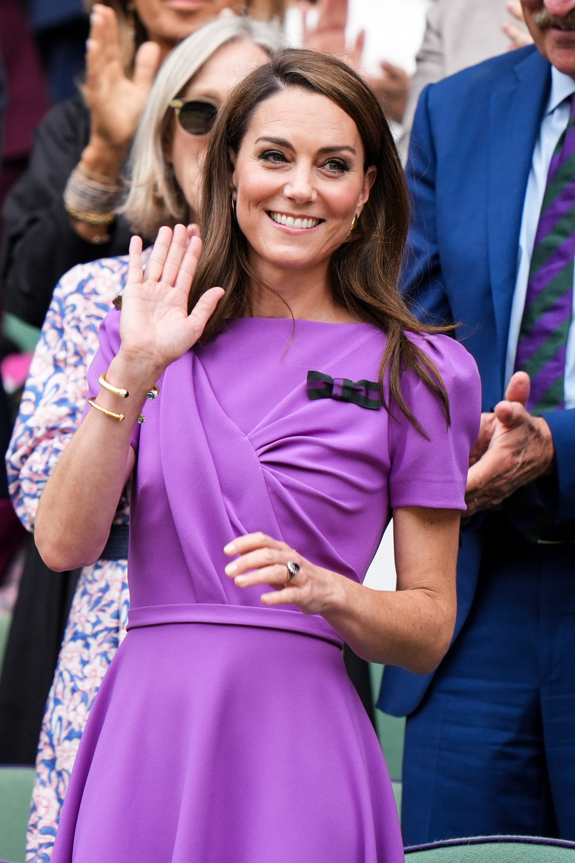 Kate Middleton, wearing a stylish purple dress with a black bow accent, waves while standing in a crowd