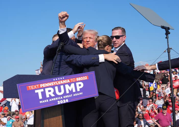 Donald Trump on stage in a suit at a rally, surrounded by security and staff, with a &quot;Trump 2024&quot; sign on the podium in front of them