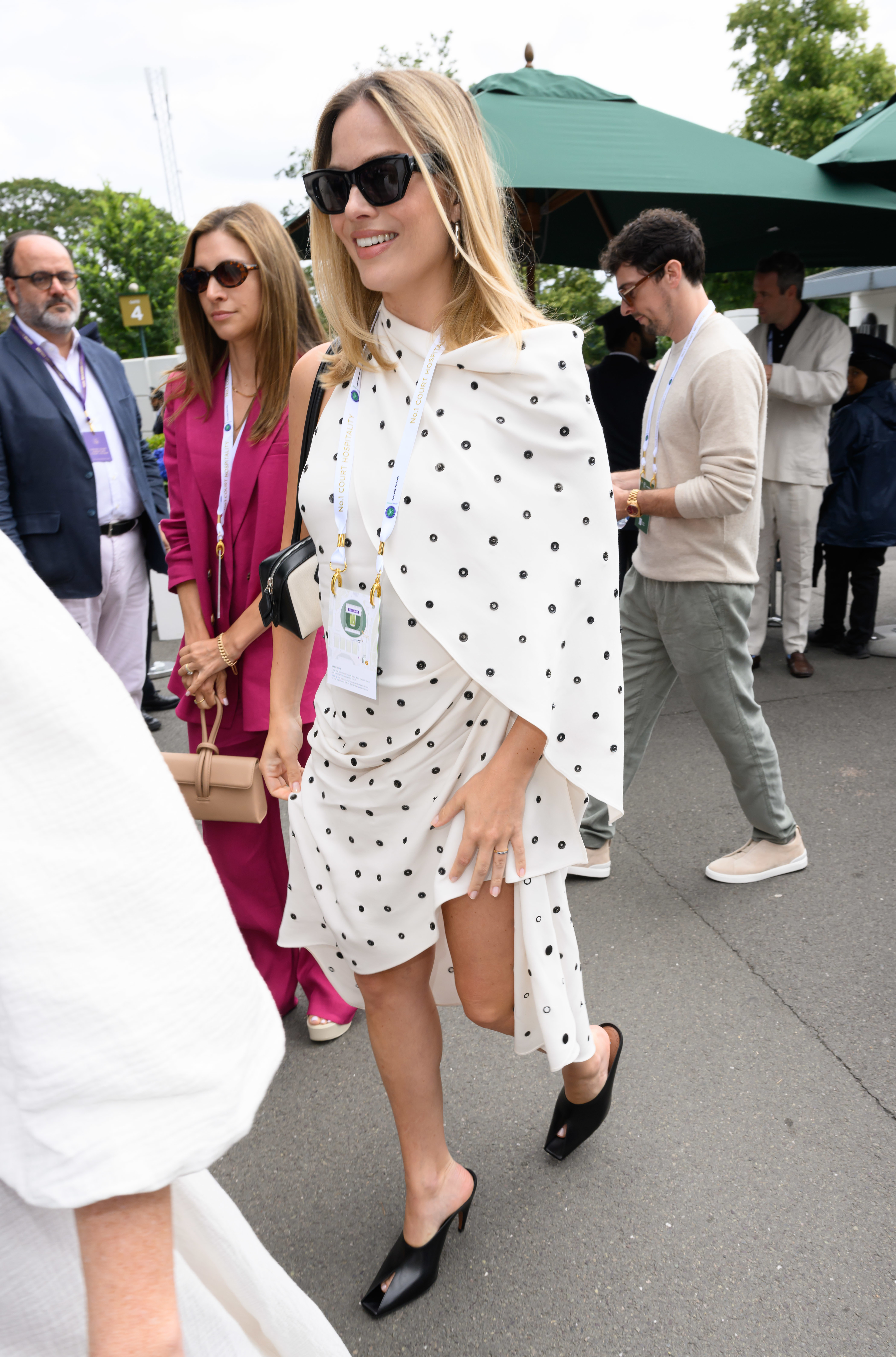 Margot Robbie walks outside in a white dress with black polka dots, wearing sunglasses and black heel shoes, among other casually dressed individuals