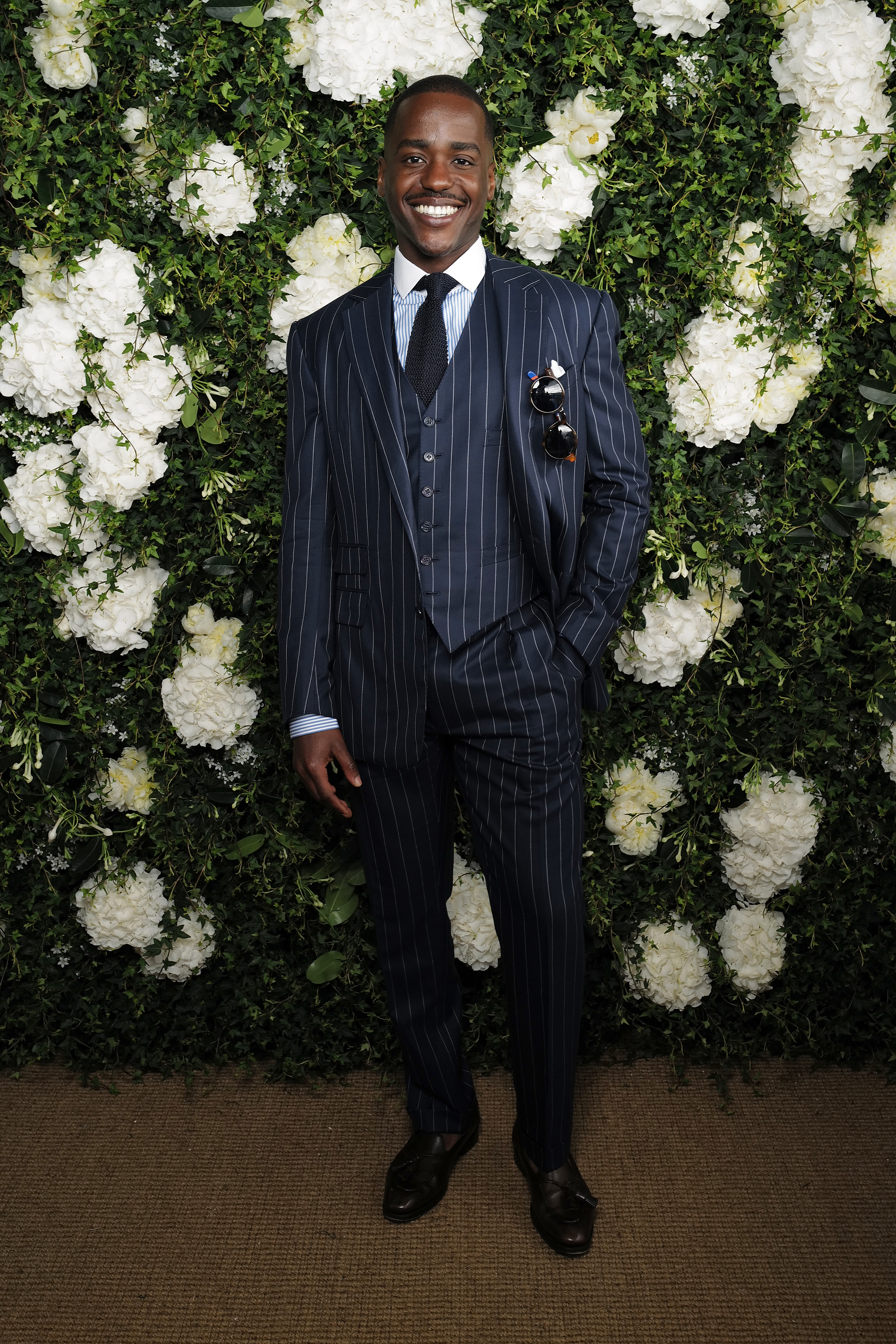 Ncuti Gatwa stands in front of a floral wall, wearing a pinstripe suit with a vest, black tie, and holding sunglasses. He is smiling with his hand in his pocket