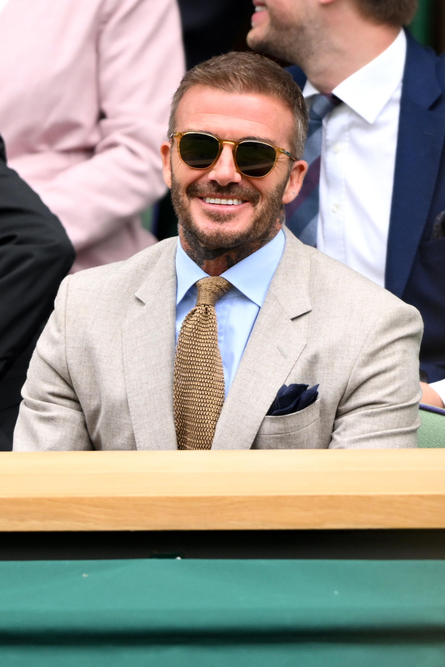 David Beckham wearing a tan suit, light blue shirt, and gold tie, seated and smiling at an event