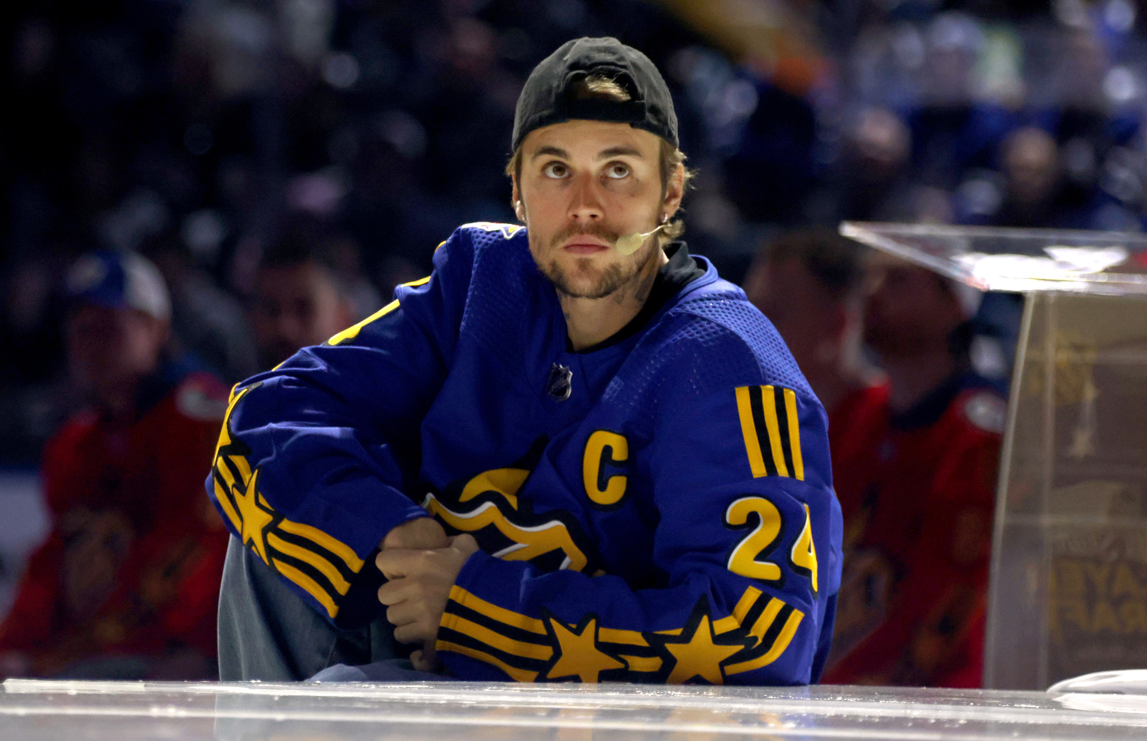Justin Bieber, wearing a sports jersey and backward cap, sits on the edge of a stage or platform during an event