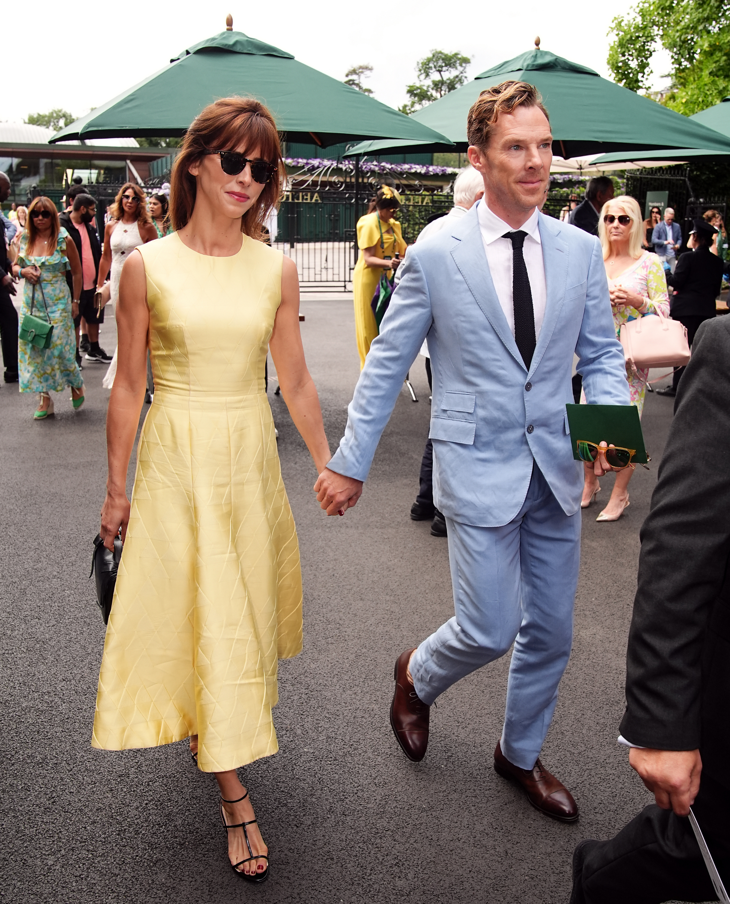 Benedict Cumberbatch in a light suit and tie, holding hands with Sophie Hunter in a sleeveless dress, surrounded by people at an outdoor event