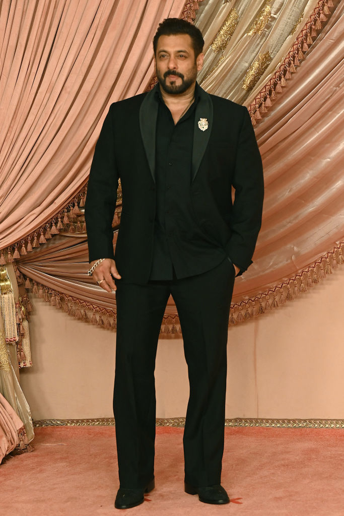 Salman Khan stands in front of an ornate curtain, wearing a well-tailored suit and dress shirt
