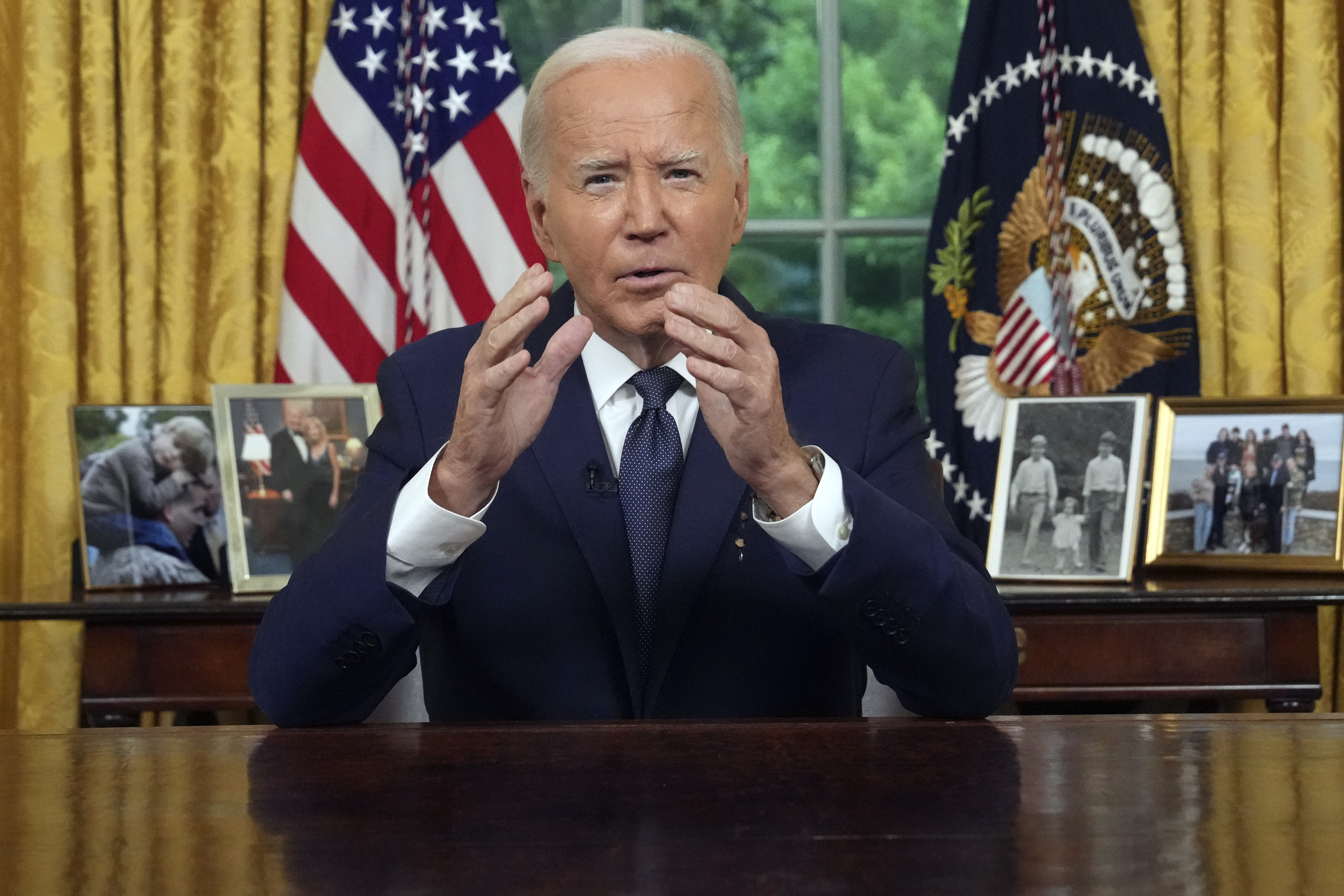 Joe Biden is seated at a desk in the Oval Office, speaking with his hands raised. Behind him are U.S. flags and framed photos