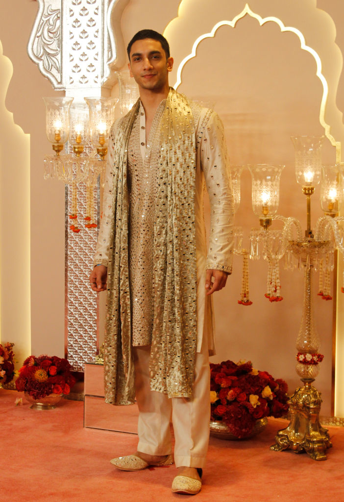 Vedang Raina in an embellished traditional outfit with a long scarf, standing indoors with ornate decorations and flowers in the background