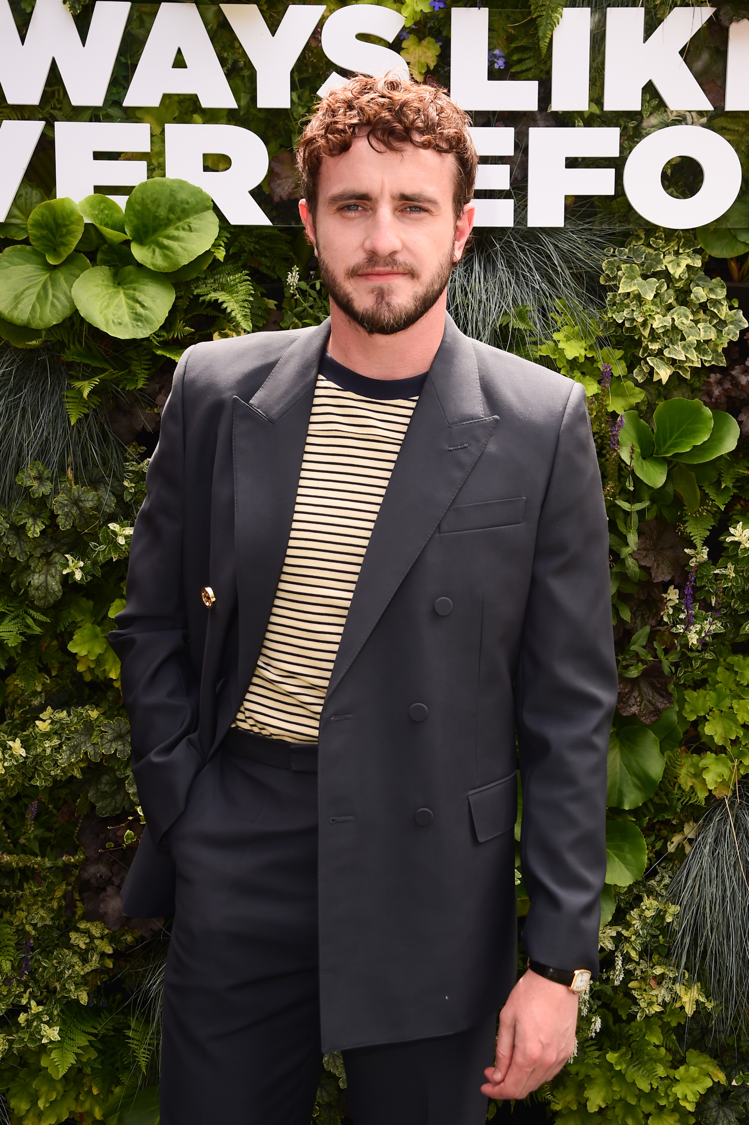 Paul Mescal poses in a black double-breasted suit with a striped shirt, standing in front of a leafy backdrop at an event