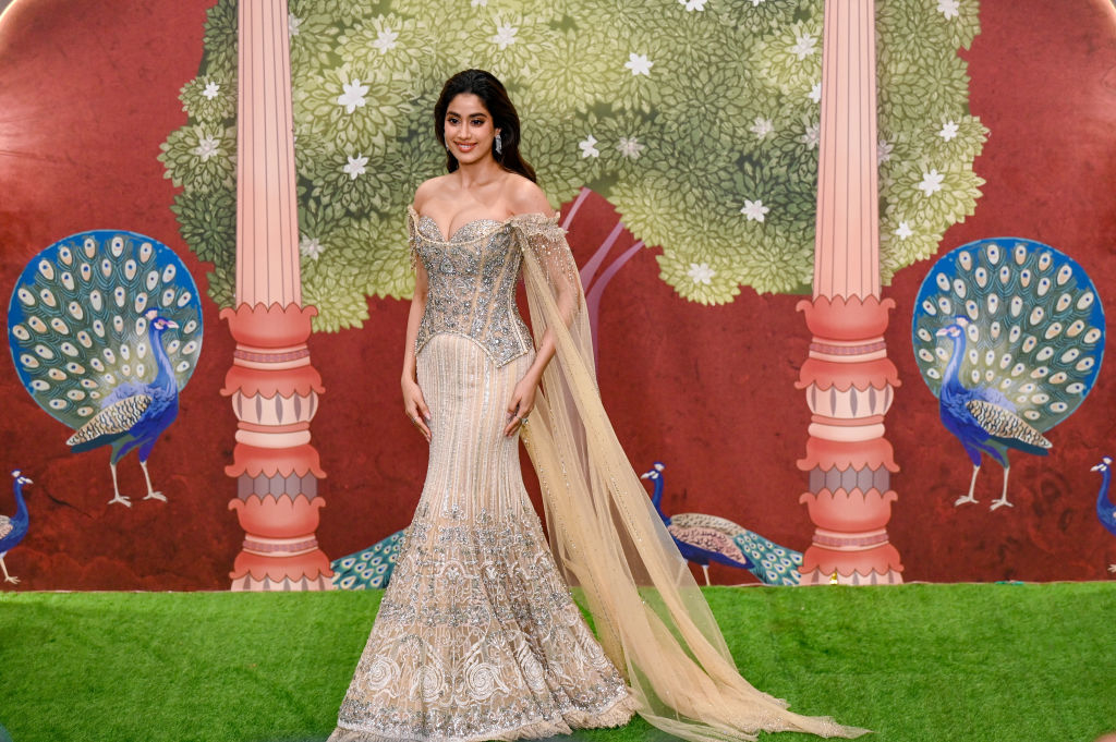 Janhvi Kapoor poses on the red carpet in an ornate, off-shoulder gown with intricate details, standing in front of a decorative backdrop featuring peacocks and trees