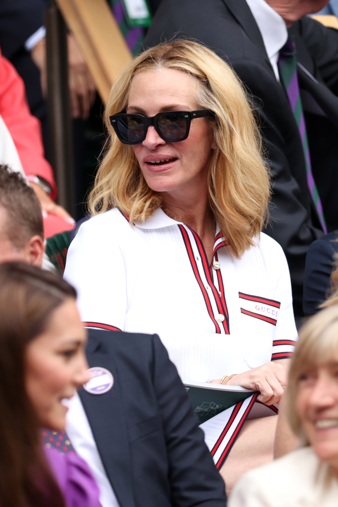 Julia Roberts is seated in a crowd, wearing a stylish white Gucci shirt with red stripes and large black sunglasses