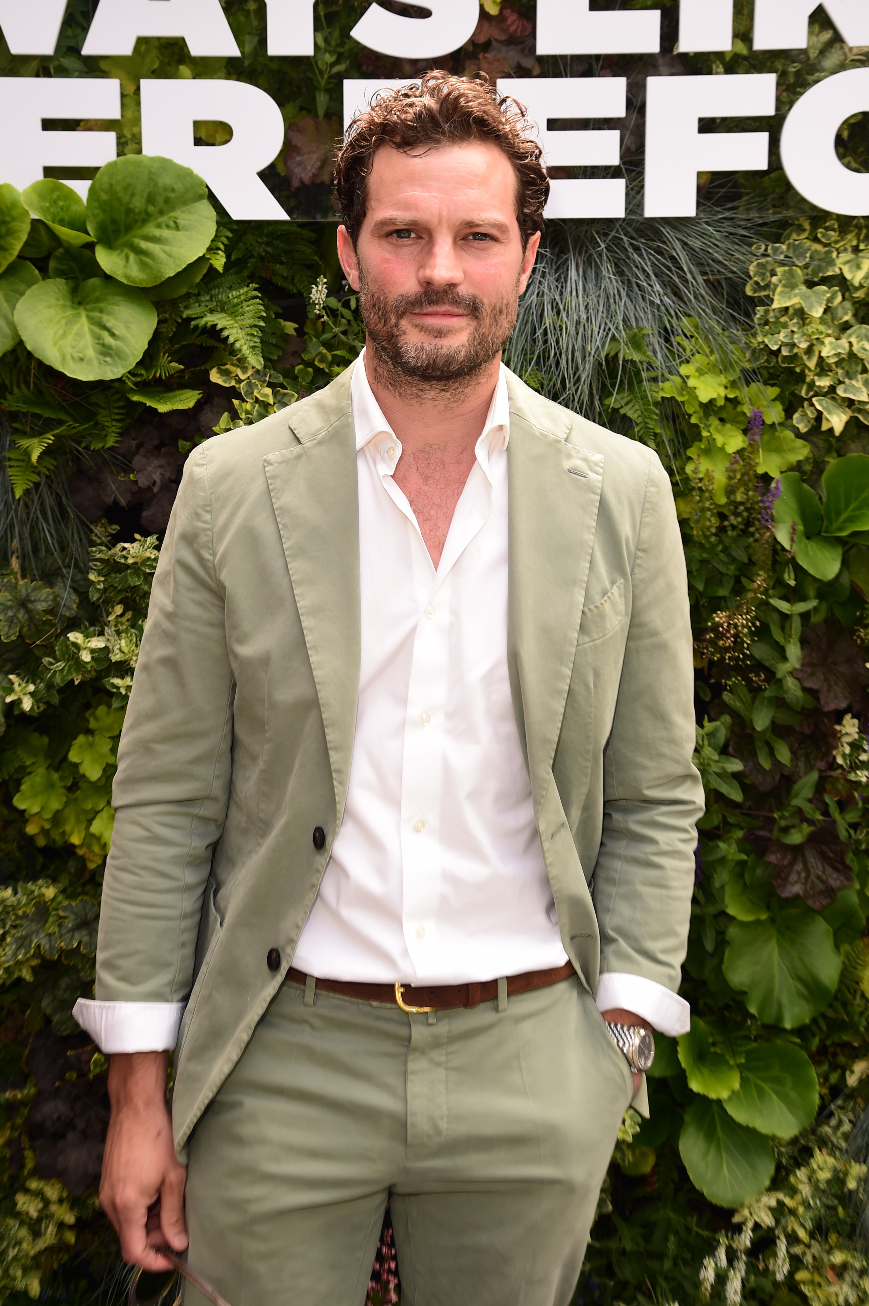 Jamie Dornan is standing in front of a plant-covered backdrop, wearing a casual suit with a white shirt