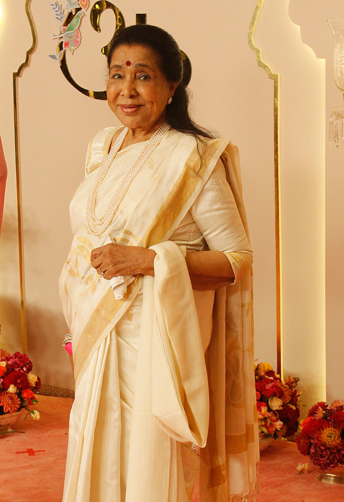 Asha Bhosle in a traditional sari with layered necklaces, standing on a red carpet