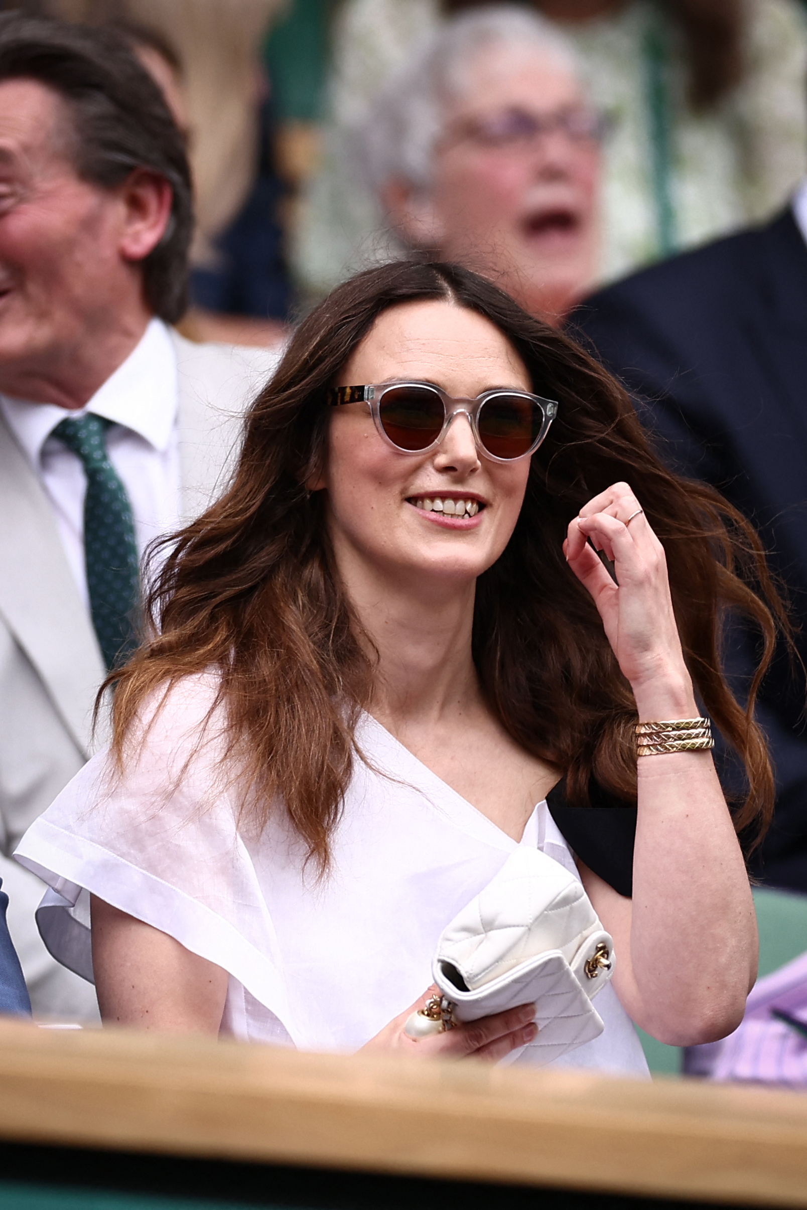 Keira Knightley is seated at an outdoor event, wearing sunglasses and a fashionable, off-the-shoulder white blouse. People in the background are watching intently