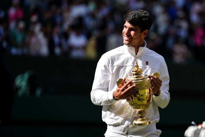 Carlos Alcaraz smiles while holding a Wimbledon trophy and wearing a white jacket and pants
