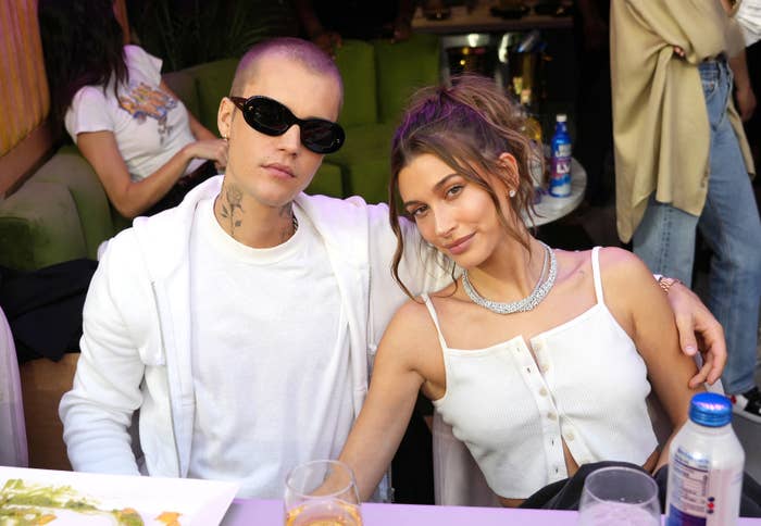 Justin Bieber, wearing sunglasses and a hoodie, sits beside Hailey Bieber, dressed in a buttoned top and necklace, at an event