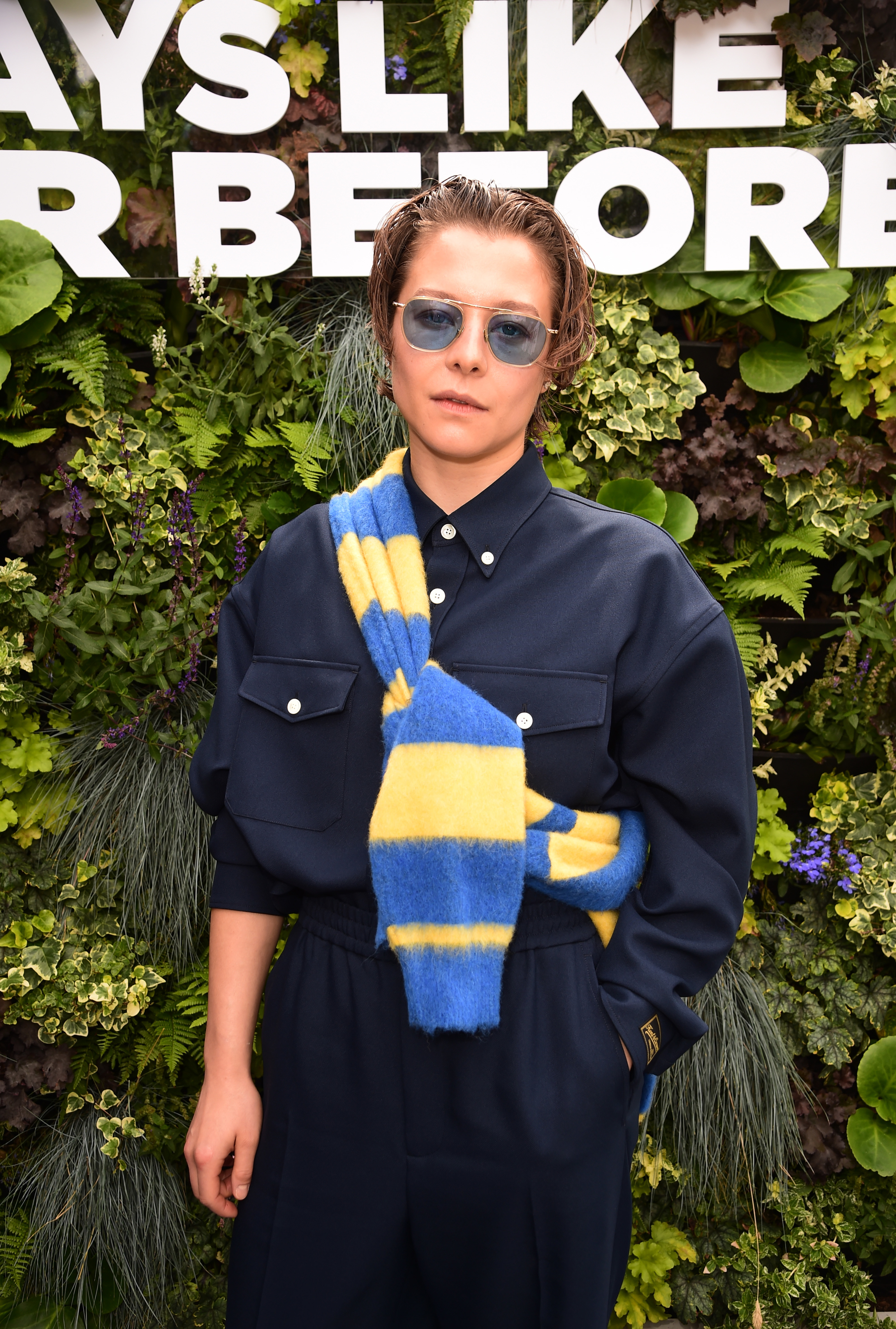 Emma D'Arcy wearing sunglasses and a dark outfit with a blue and yellow scarf stands in front of a leafy wall under a partially visible sign