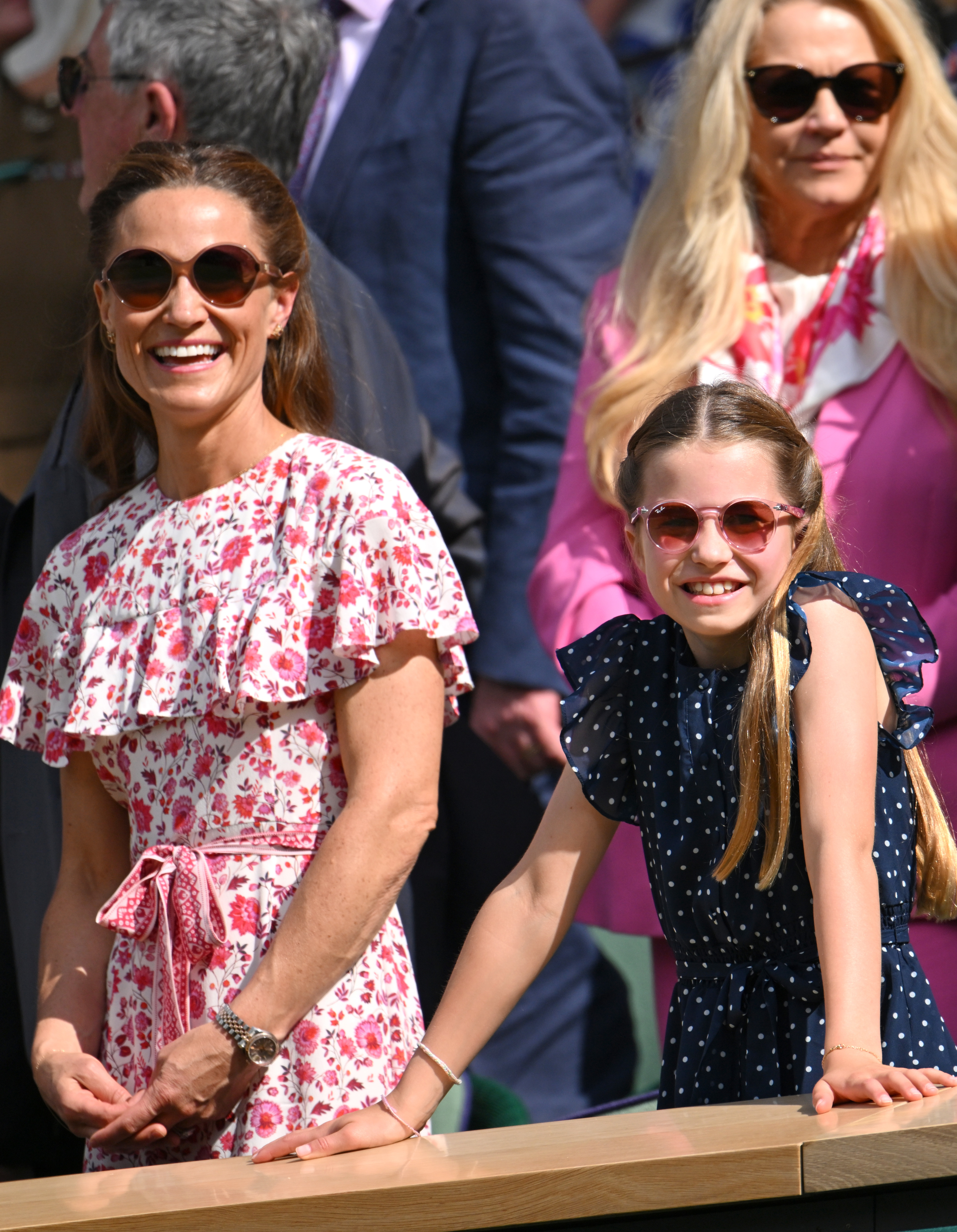 Pippa Middleton and her daughter smile while watching an outdoor event. Both wear sunglasses, with Pippa in a floral dress and her daughter in a polka-dot dress