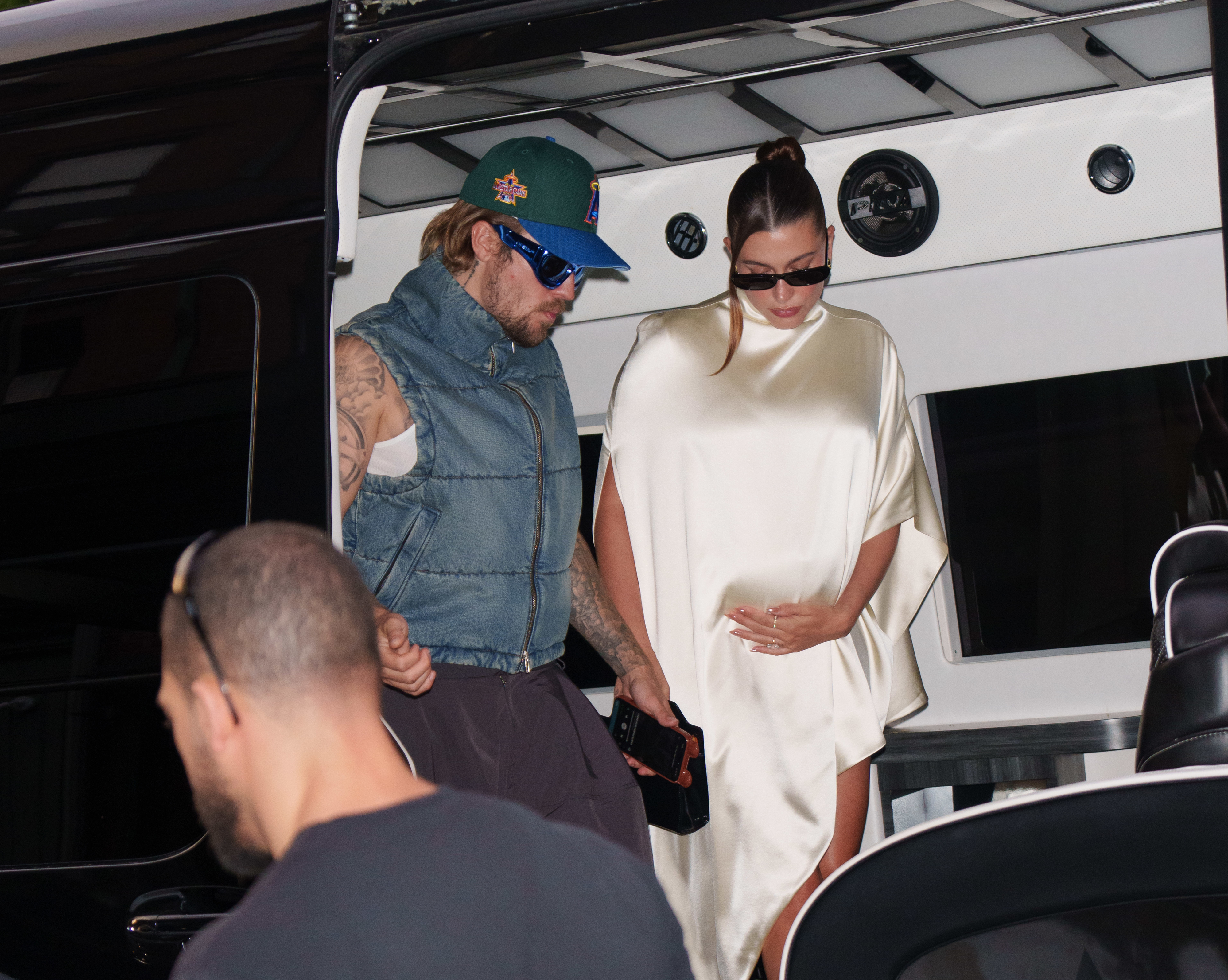 Justin Bieber in a denim vest and cap, and Hailey Bieber in a silk dress and sunglasses, step out of a vehicle