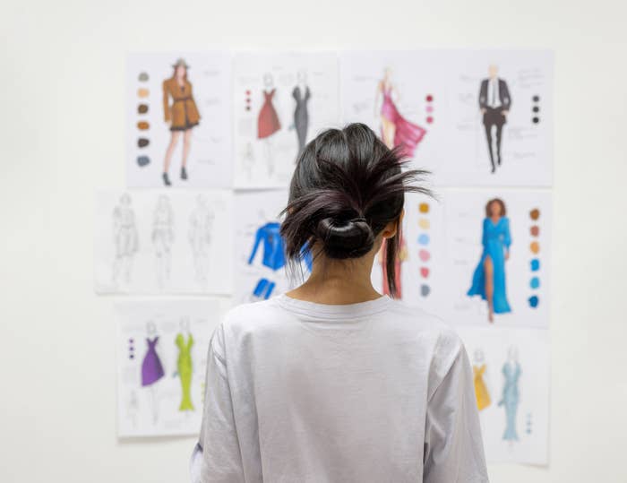 A person with their back turned is looking at a wall with multiple fashion sketches and color swatches. The person is wearing a white shirt. No names are visible
