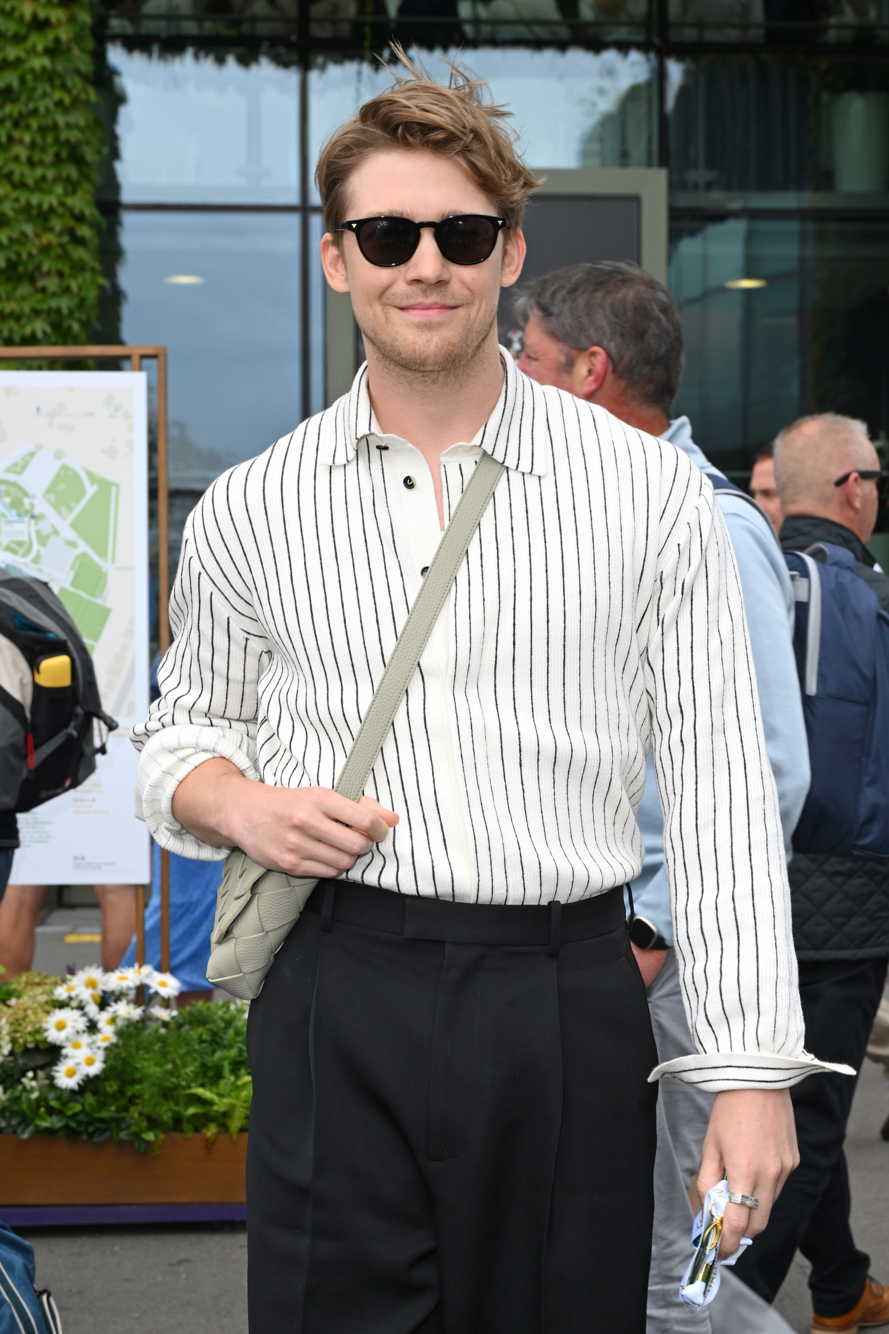 Joe Alwyn is smiling, wearing a striped sweater, black pants, sunglasses, and a crossbody bag, while holding an item in his hand. He is standing outdoors