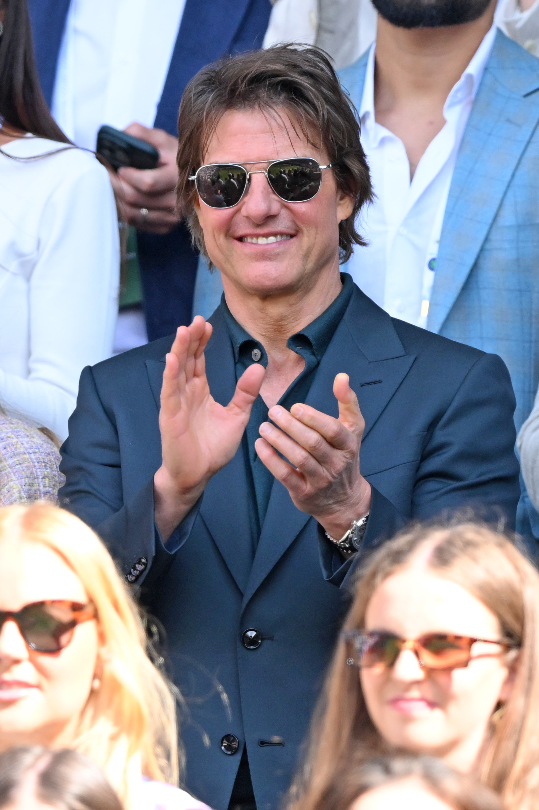 Tom Cruise, in a sleek suit with sunglasses, clapping at an outdoor event surrounded by others