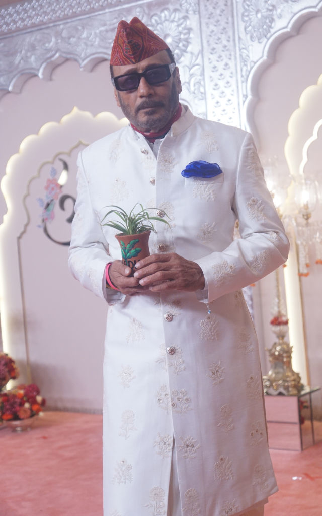 Jackie Shroff stands on a red carpet in an ornate sherwani, holding a small potted plant; he wears sunglasses a patterned hat