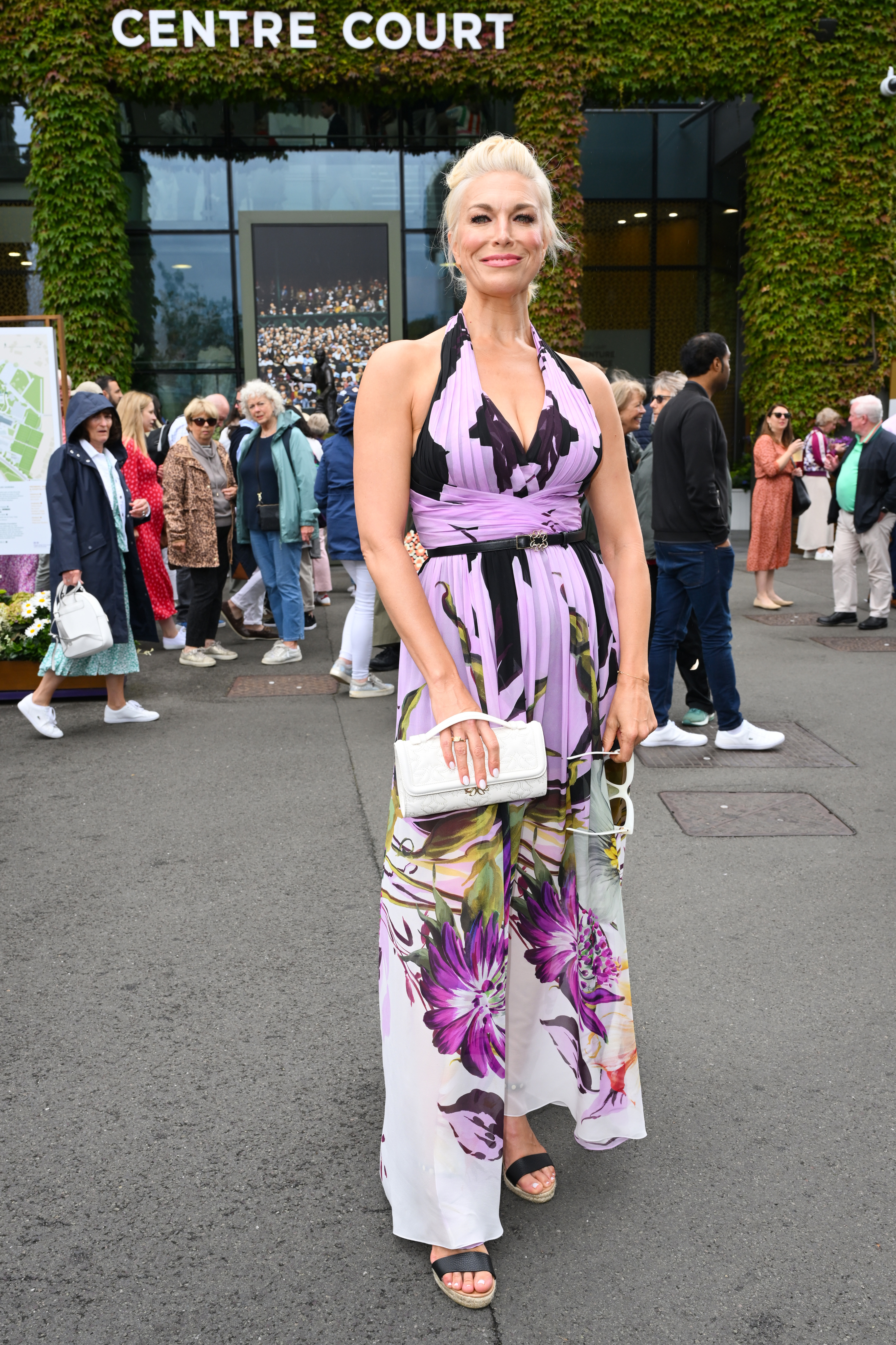 Hannah Waddingham dressed in a floral halter-neck jumpsuit, holding a white clutch, stands outside Centre Court with a crowd in the background