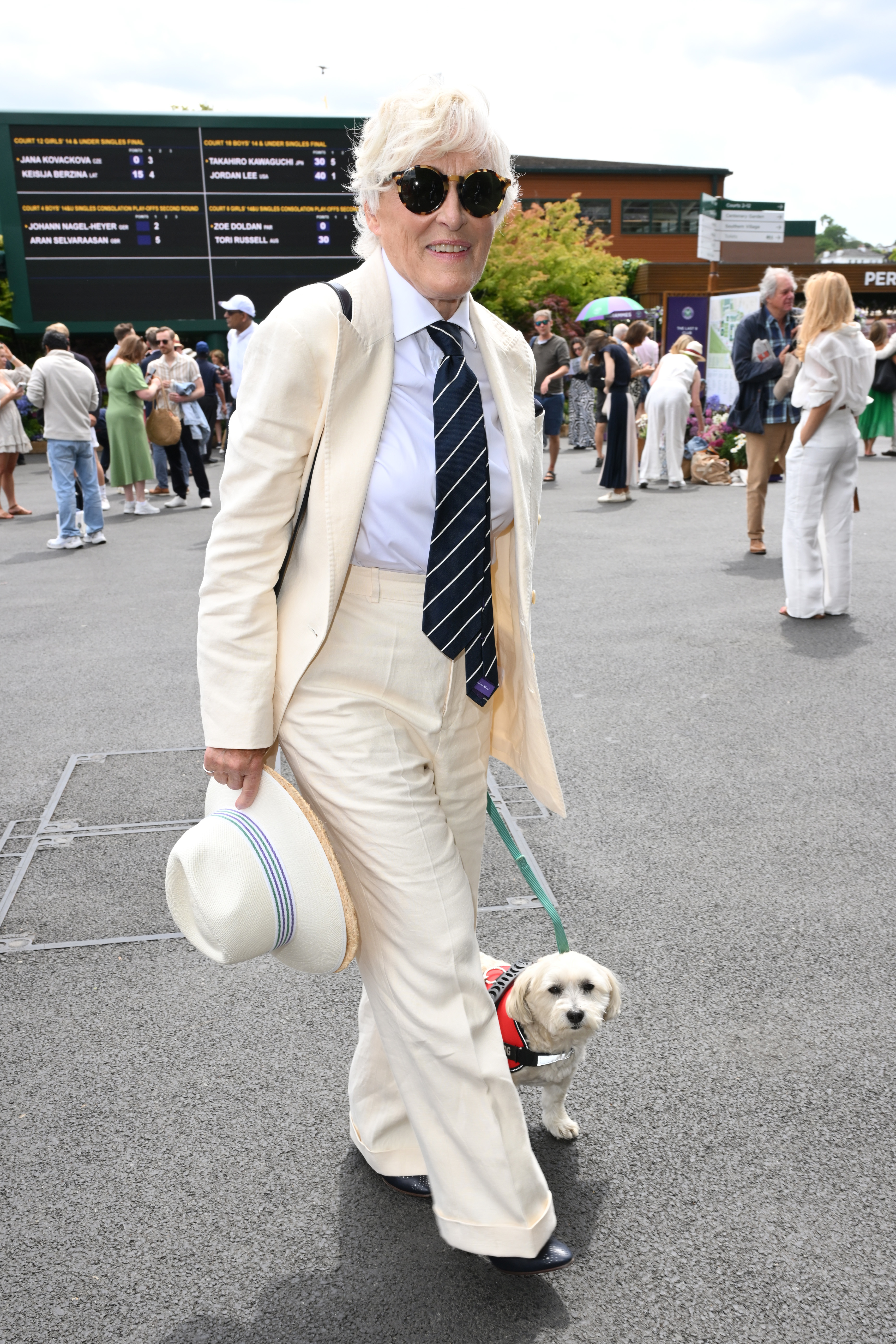 Glenn Close in a classic cream suit with a tie and hat, walking a small dog in a red harness at an outdoor event