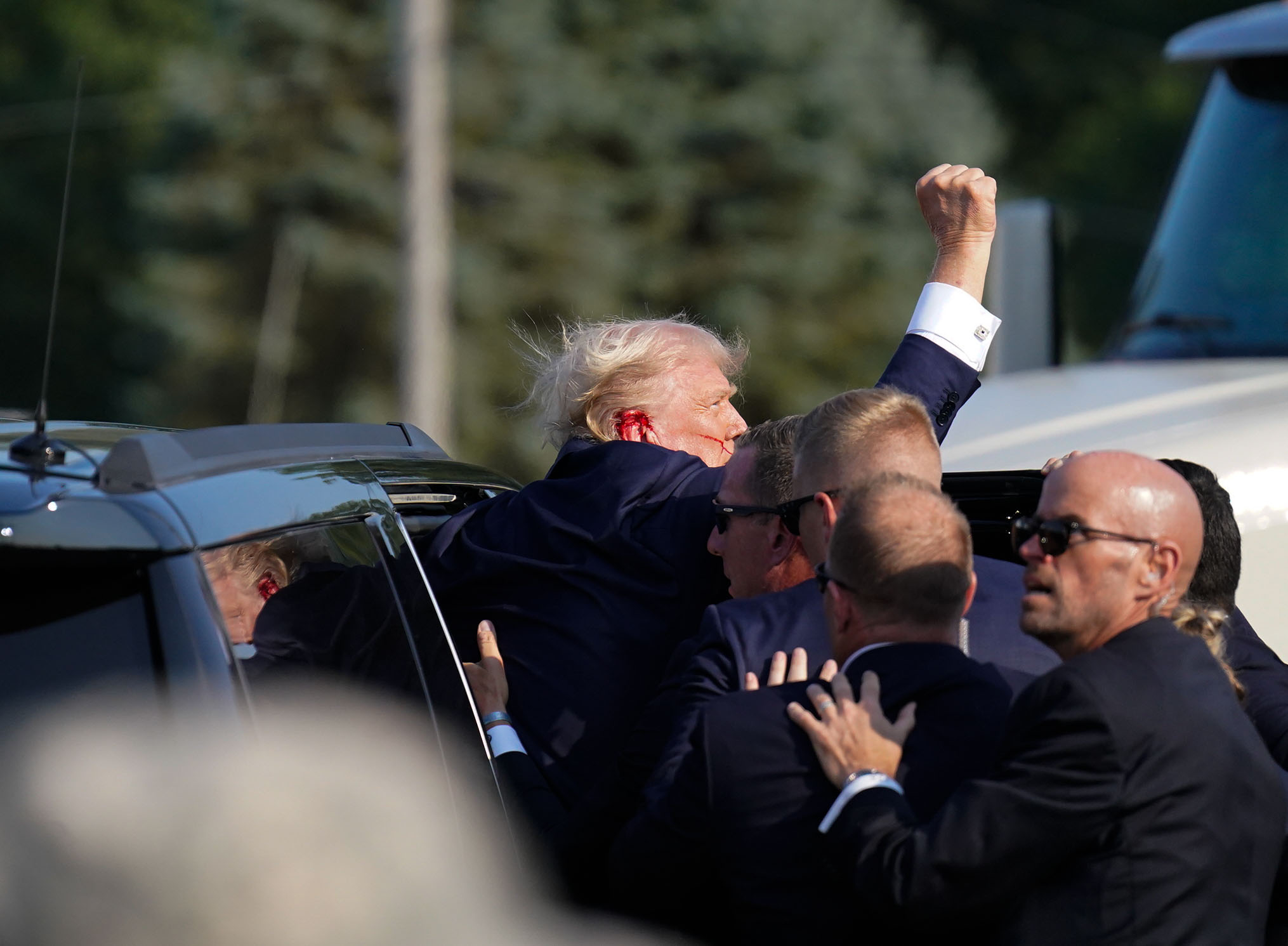 Donald Trump raises his fist in the air while being surrounded by multiple security personnel outdoors