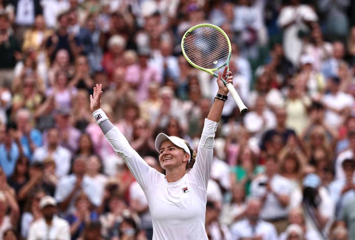Barbora Krejcikova celebrates with a tennis racket while wearing sports attire on a tennis court, with a crowd cheering in the background