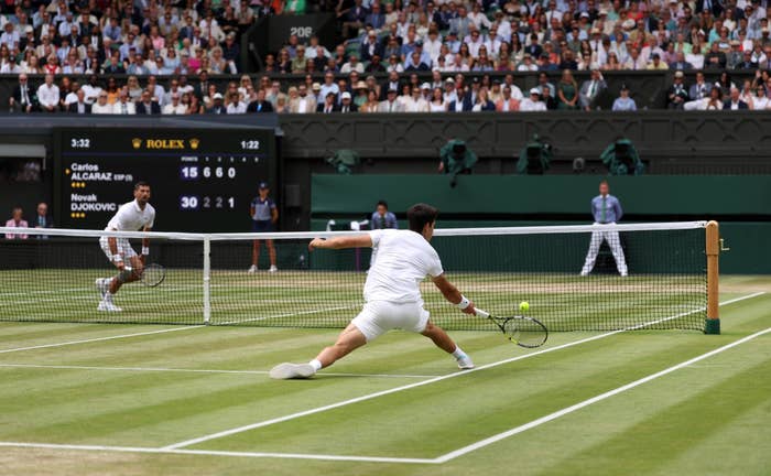 Carlos Alcaraz and Novak Djokovic playing at Wimbledon, mid-action on a grass court with a crowd and scoreboard in the background