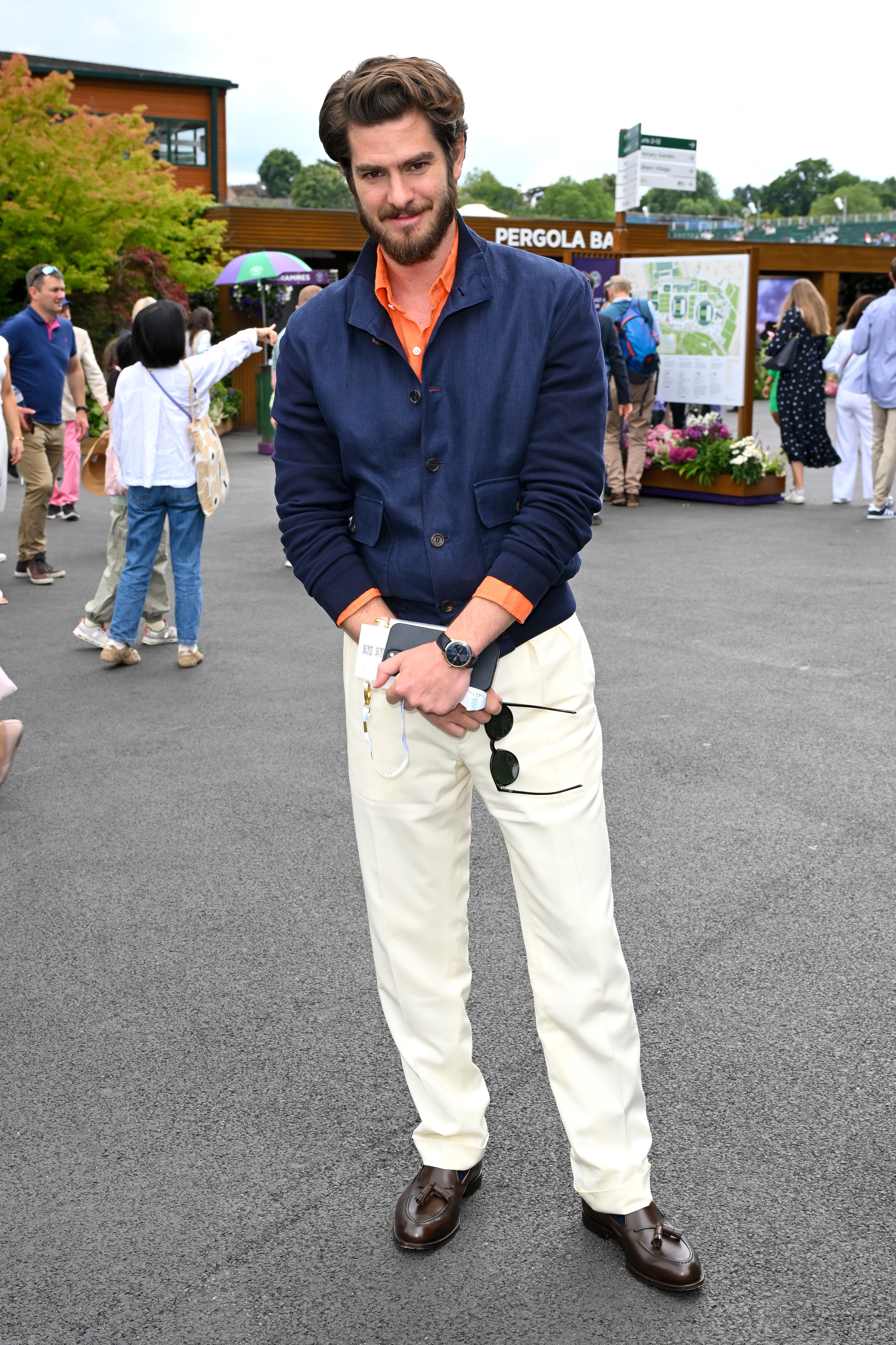 Andrew Garfield poses outdoors, wearing a navy blazer, orange shirt, and white pants, holding sunglasses and tickets. People and greenery are in the background
