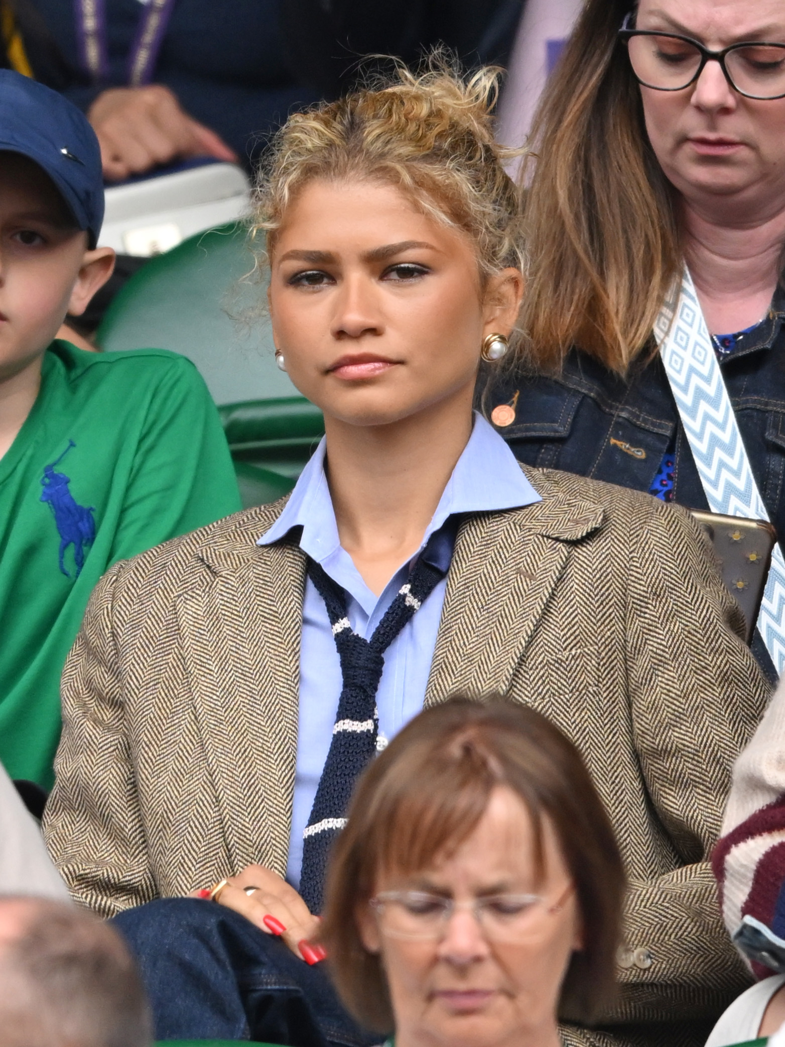 Zendaya sitting in an audience, wearing a blazer, blue shirt, and tie, with her hair tied up. She is surrounded by other attendees