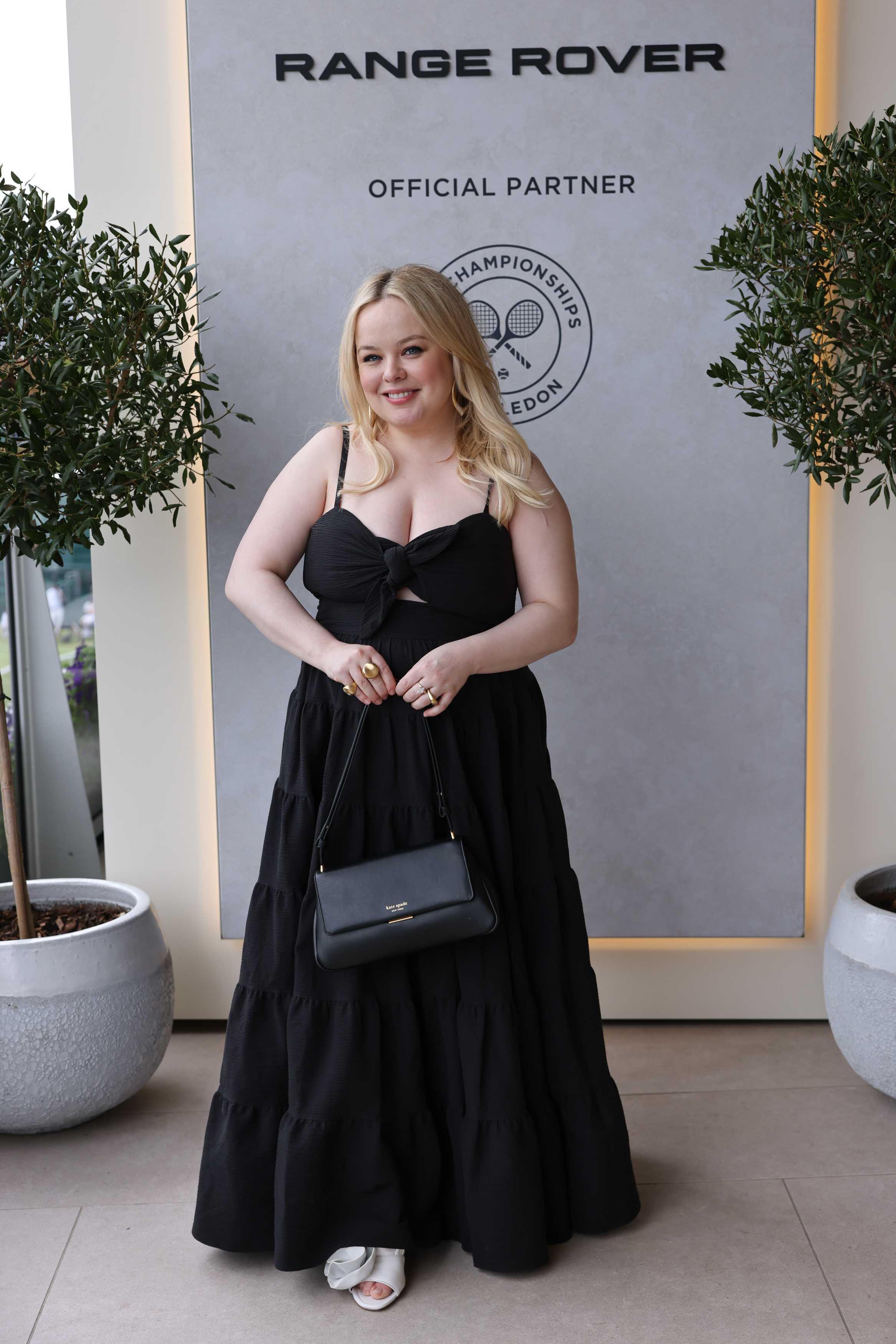 Nicola Coughlan in a long black strapless dress, holding a black purse, poses in front of a Range Rover and Wimbledon branded backdrop