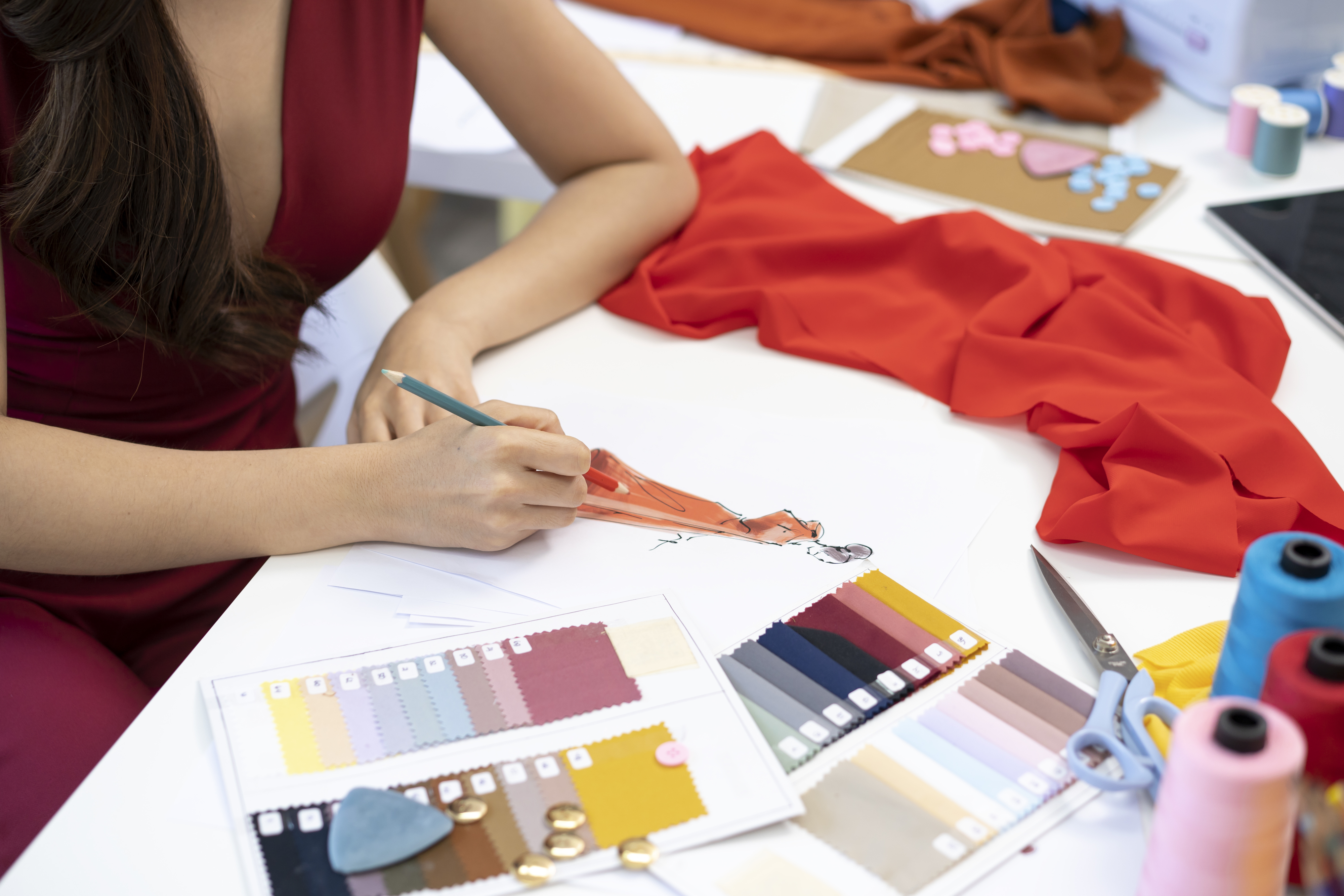 A person is sketching a dress while surrounded by fabric swatches, sewing supplies, and threads on a work table in a design studio