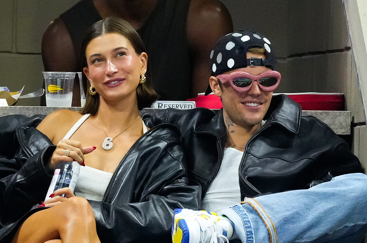 Hailey Bieber and Justin Bieber sit together in an audience. Hailey wears a white outfit and Justin wears a black leather jacket, beanie, and sunglasses