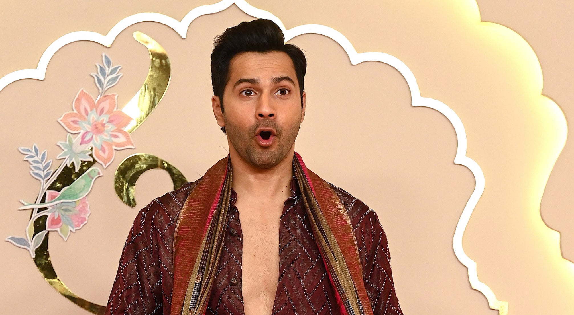 Varun Dhawan in a maroon patterned kurta, white pants, and a multicolored scarf at an event, standing in front of floral arrangements