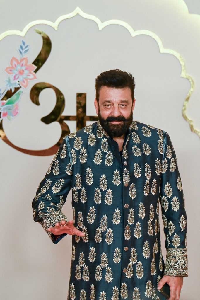 Sanjay Dutt wearing an elaborate, patterned traditional outfit at a public event. He is smiling and posing for the cameras