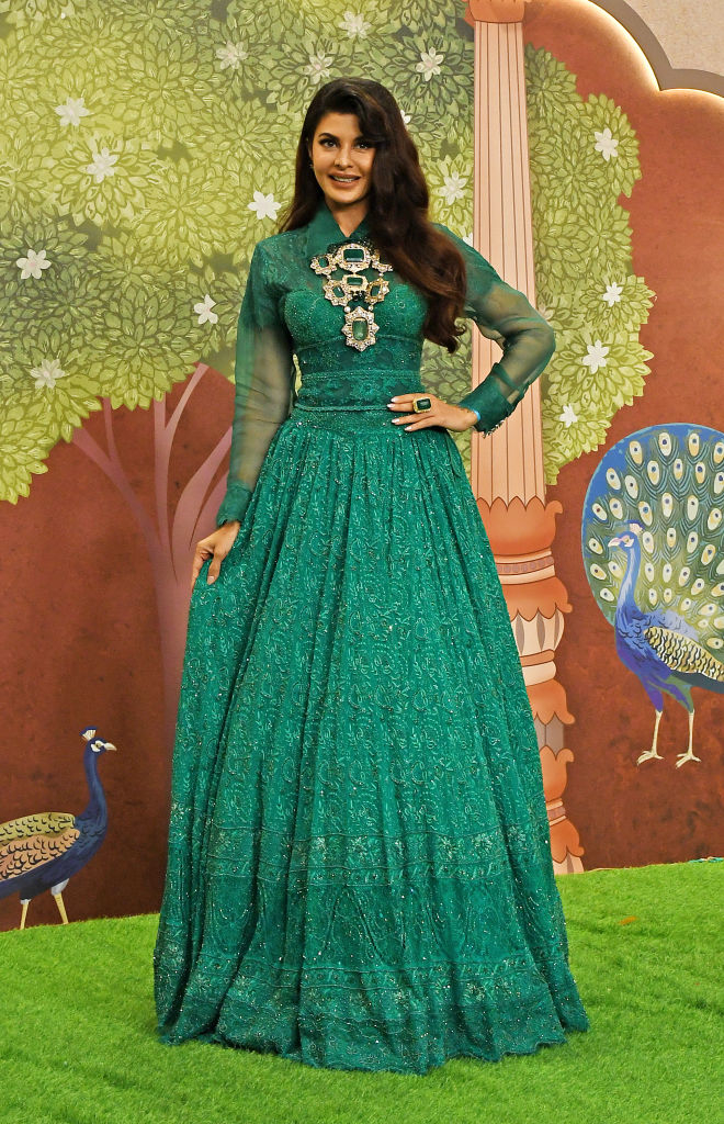 Jacqueline Fernandez poses outdoors in an intricately designed long gown with sheer sleeves, in front of a backdrop featuring trees and peacocks